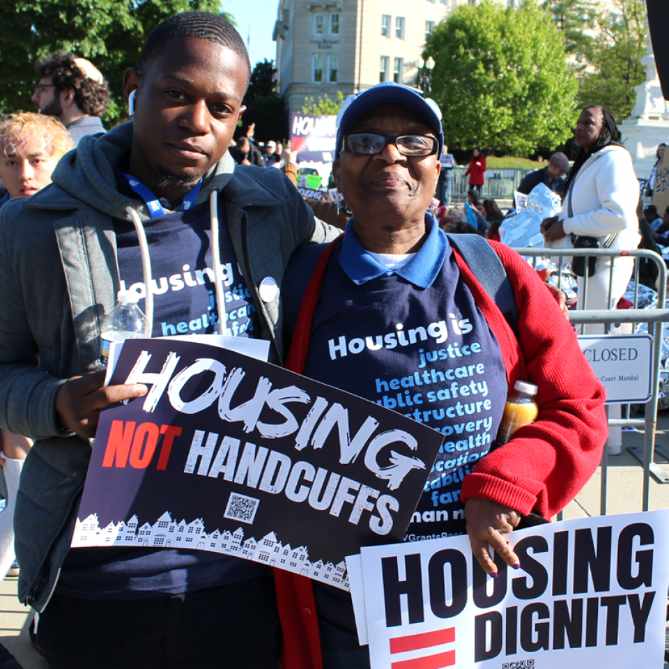 Two people at a rally hold signs that say "Housing not handcuffs" and "Housing = dignity"