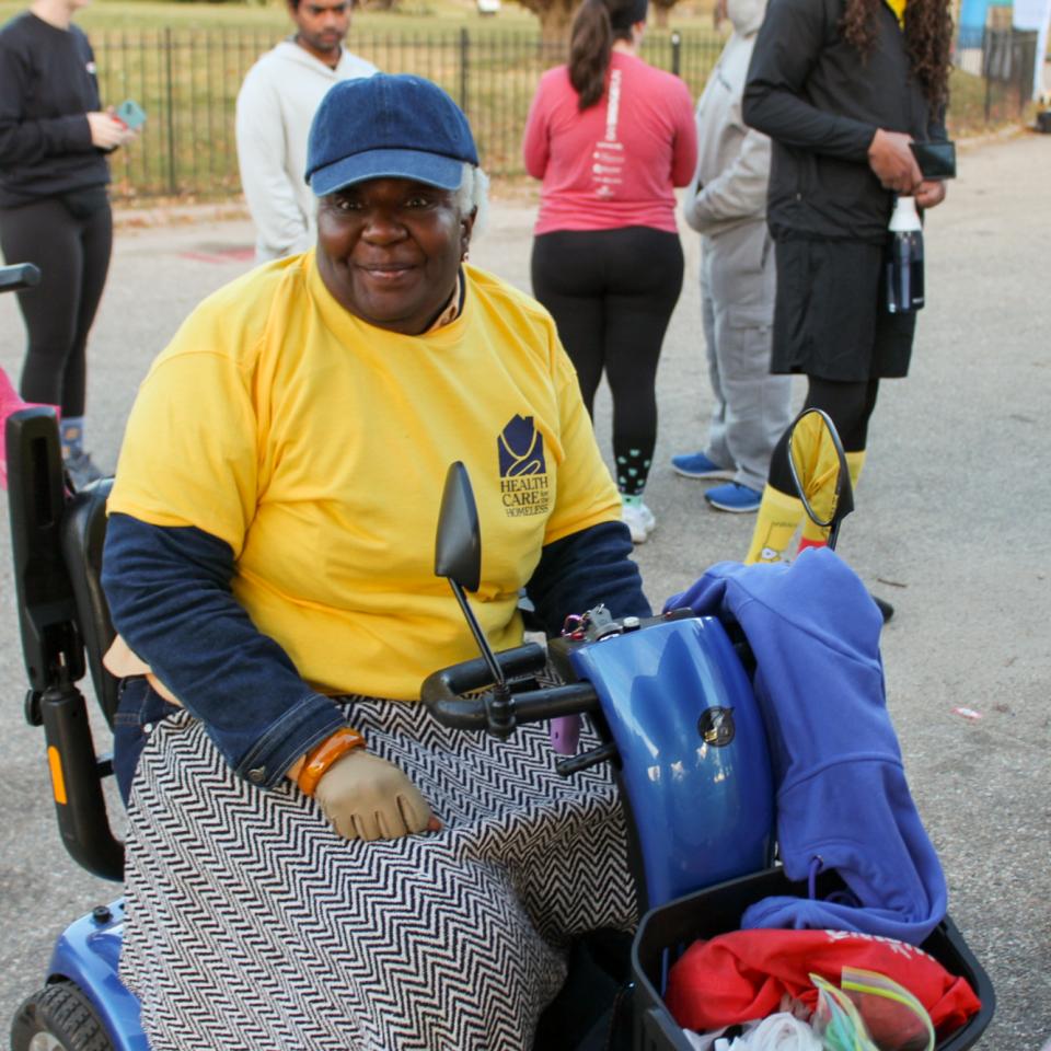 A dark-skinned woman smiles at the camera. She's wearing a blue baseball cap and a yellow t-shirt with the Health Care for the Homeless logo.