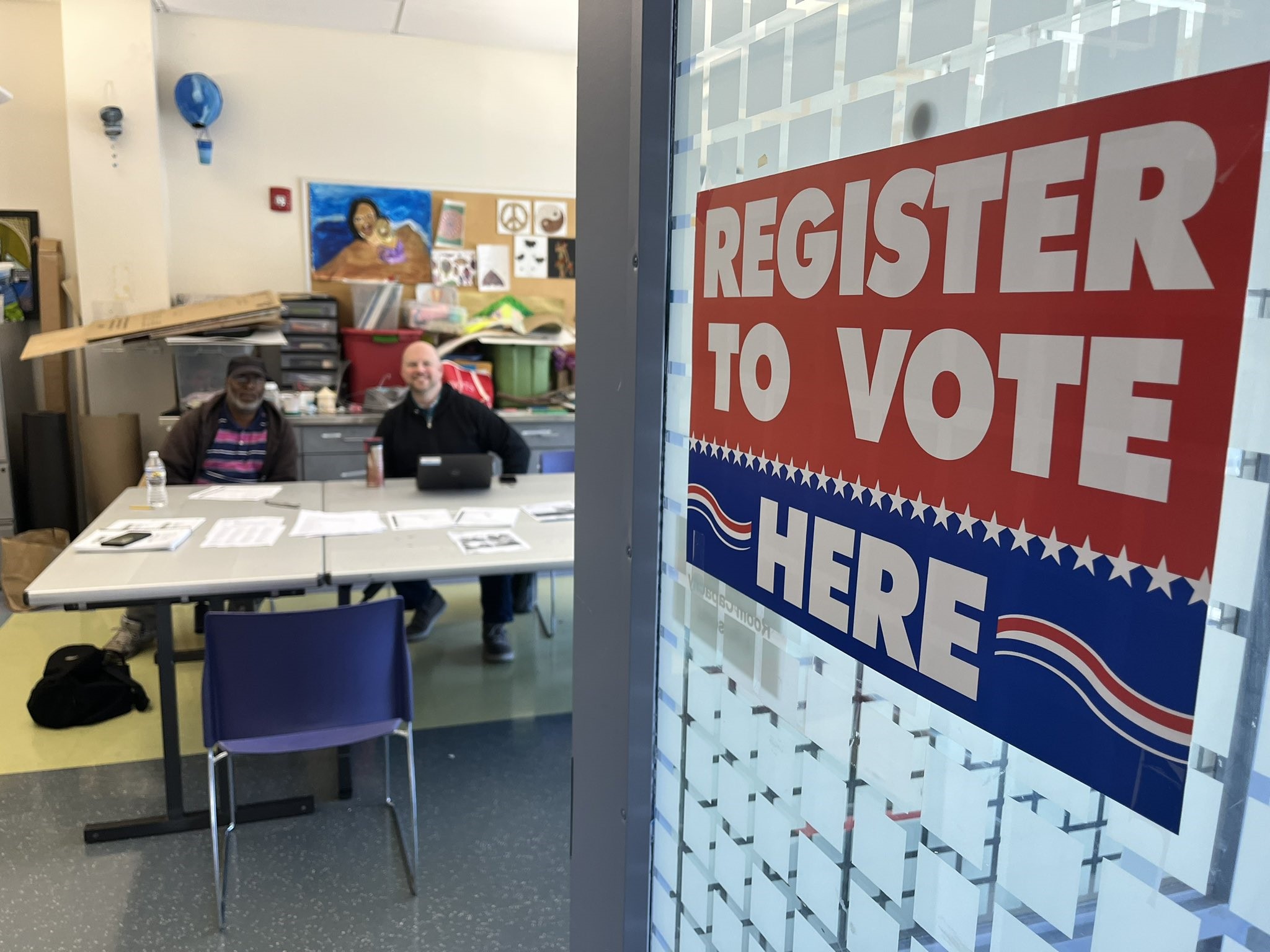 Two volunteers smile behind a desk. Sign reads "Register to vote here"